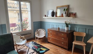 Ein Zimmer mit einem Holzkleiderschrank, Korbstühlen, einem kleinen Tisch mit farbigem geometrischen Muster, einem Fenster mit Blick auf einen Balkon und einem Regal mit Dekoration.