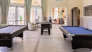 Une table de billard, une table de baby-foot et un canapé dans une pièce bien éclairée, dotée de grandes fenêtres et offrant une vue sur l’extérieur.