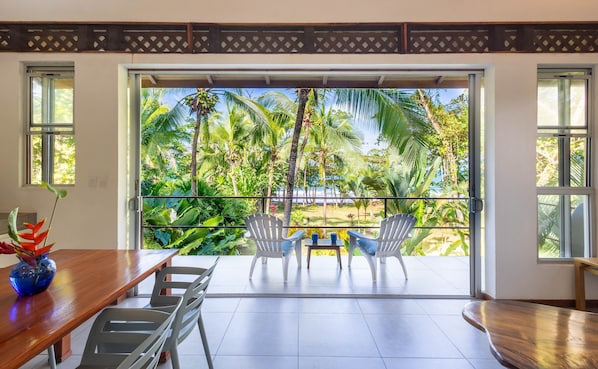Une pièce dotée d’une porte-fenêtre coulissante donnant sur un balcon aménagé avec des chaises et une table, d’où l’on peut admirer un jardin tropical et une plage.
