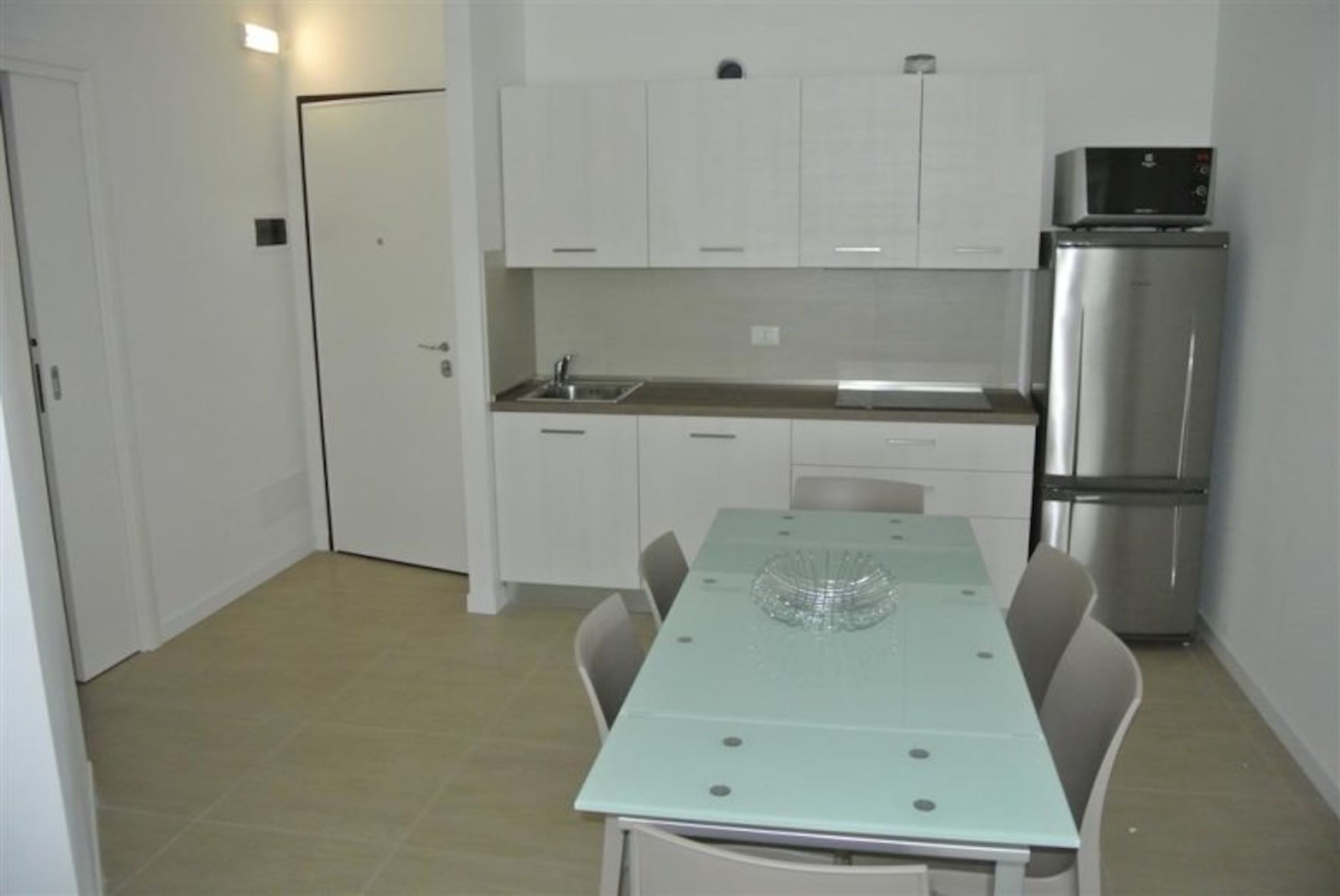A compact kitchen with white cabinets, a stainless steel refrigerator, a glass-top dining table, and a tiled floor.