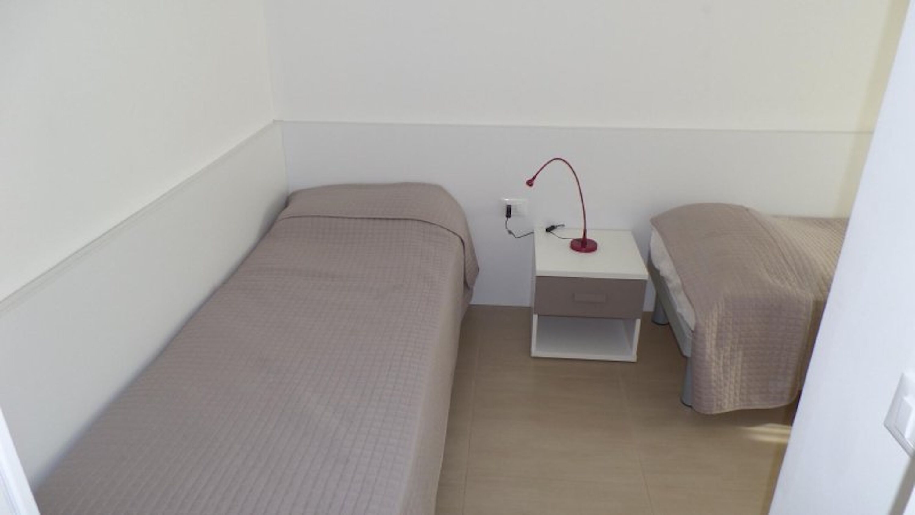 A small, single-room with two beds, a nightstand, and a desk with a lamp.