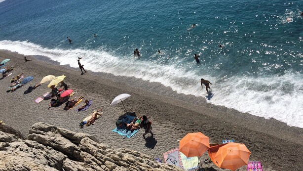 Ein Strand mit badenden Personen, Sonnenschirmen und einer felsigen Küstenlinie.