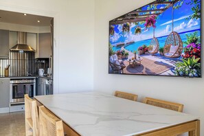 Ein Essbereich mit einem Marmortisch und Stühlen, eine Küche mit modernen Geräten und ein großes Gemälde einer tropischen Strandlandschaft.
