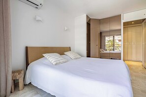 Ein Schlafzimmer mit einem großen Bett, einem Holz-Kopfteil, einem kleinen Hocker, einer weißen Wand und einer Tür, die auf einen Balkon führt.