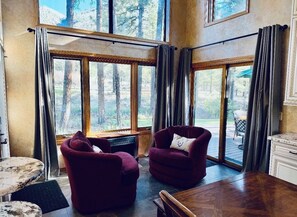 Ein Zimmer mit großen Fenstern, einem Holzespeisetisch, zwei Sesseln und Blick auf Bäume.