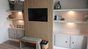 A living room with a TV mounted on a wooden panel wall, a bench, shelves with books and decorative items, and a small table with a basket.