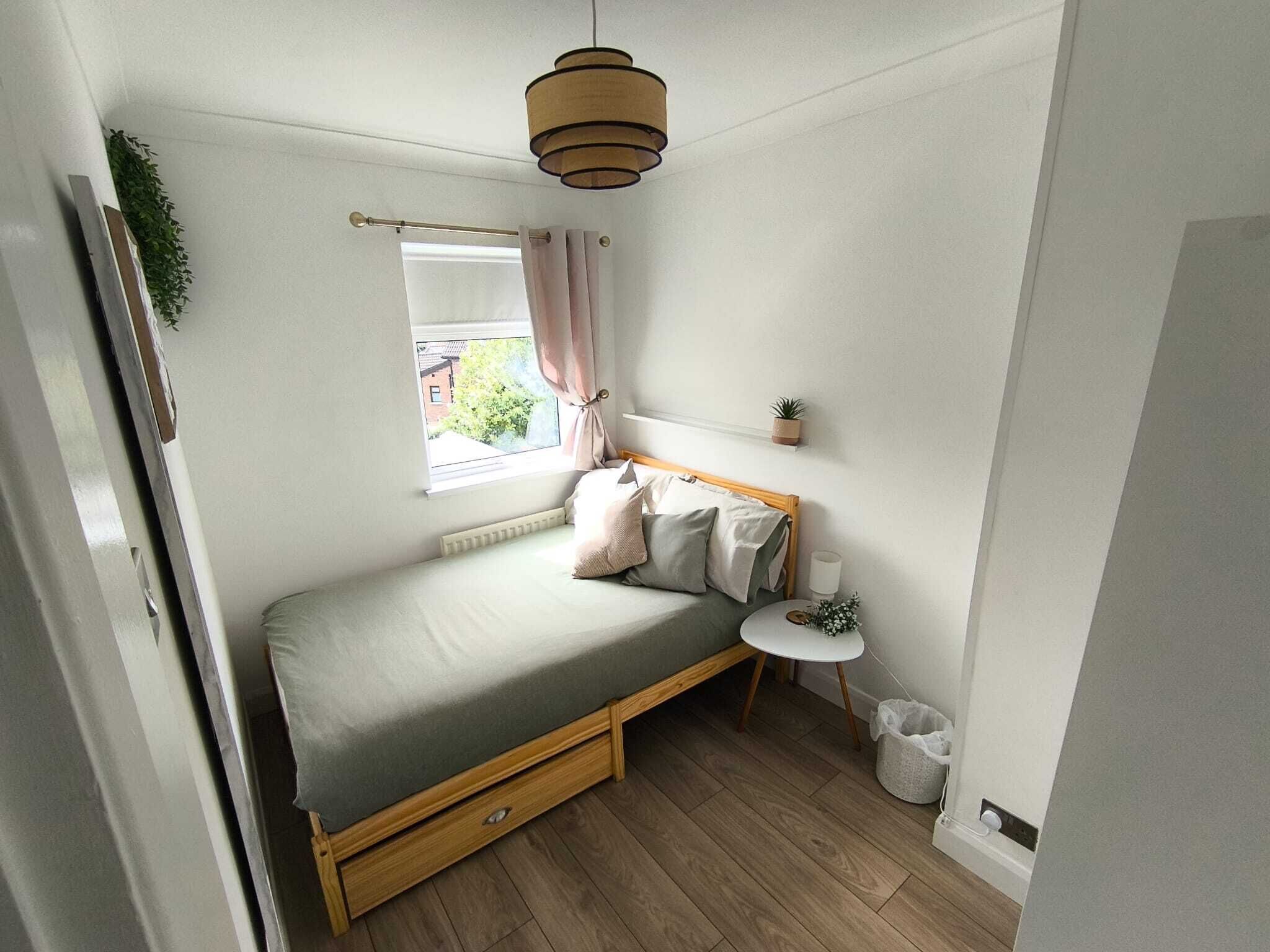 Een kleine slaapkamer met een bed, nachtkastje en een klein raam met gordijnen.