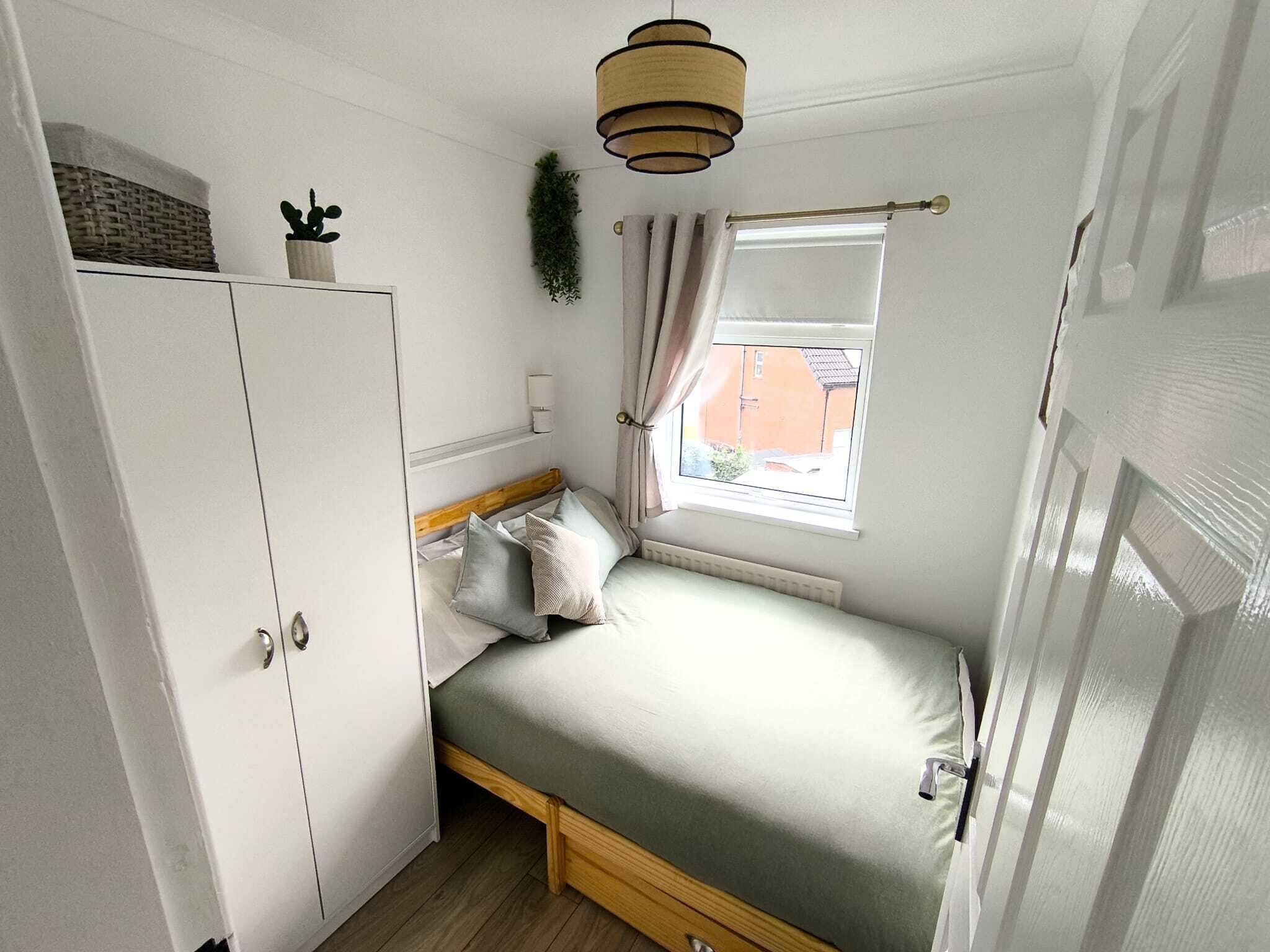A compact bedroom with a bed, a wardrobe, a window with curtains, and a hanging plant.