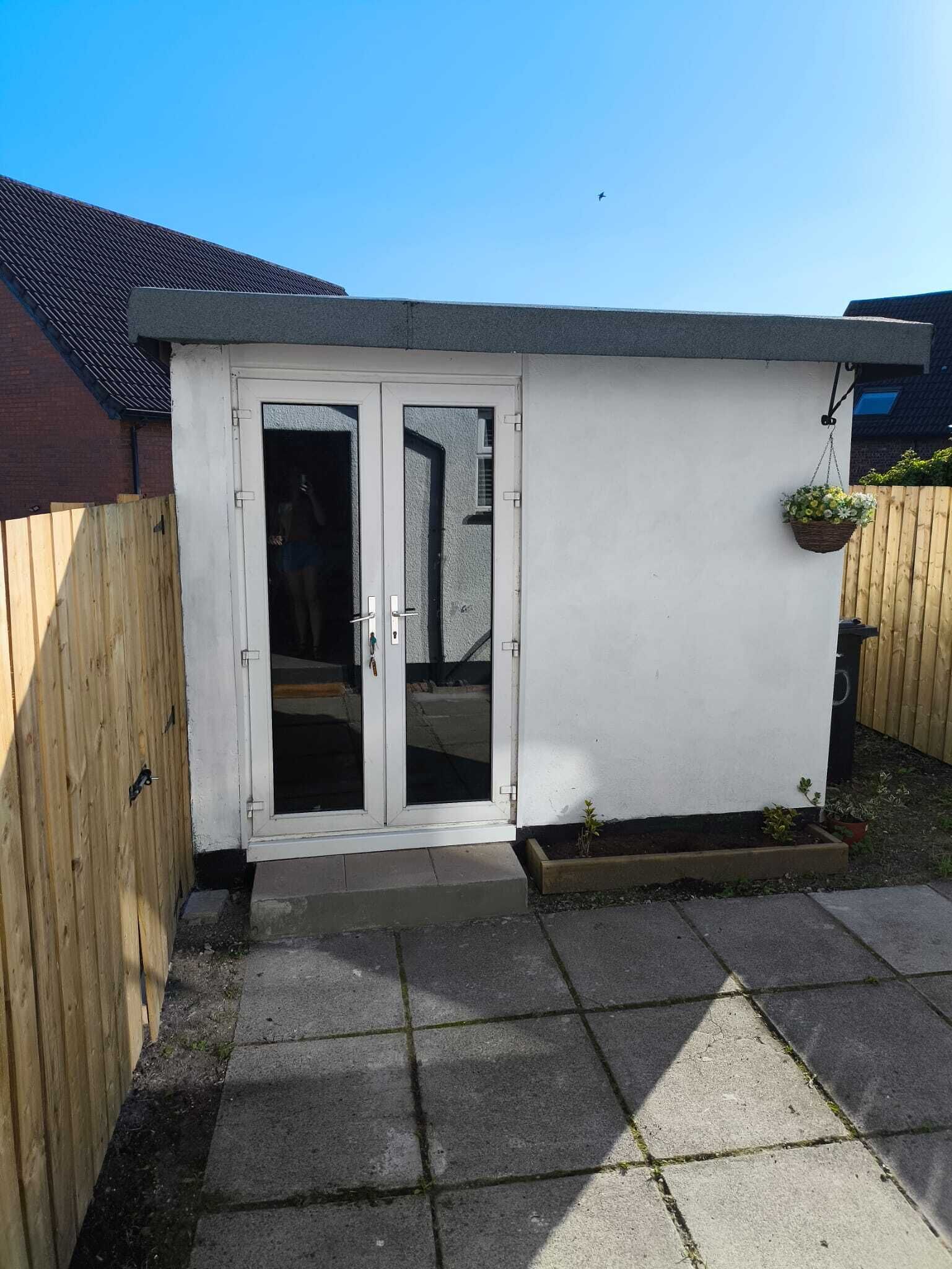 A small, enclosed patio area with a white exterior wall, a sliding glass door, and a wooden fence.
