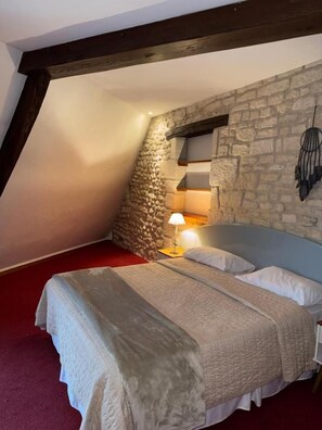 Une chambre avec un mur en pierre, un plafond en pente, un lit avec une tête de lit et une lampe de chevet.