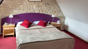 Un dormitorio con pared de piedra, una cama con cabecera morada, dos mesitas de noche con lámparas y un cuadro sobre la cama.