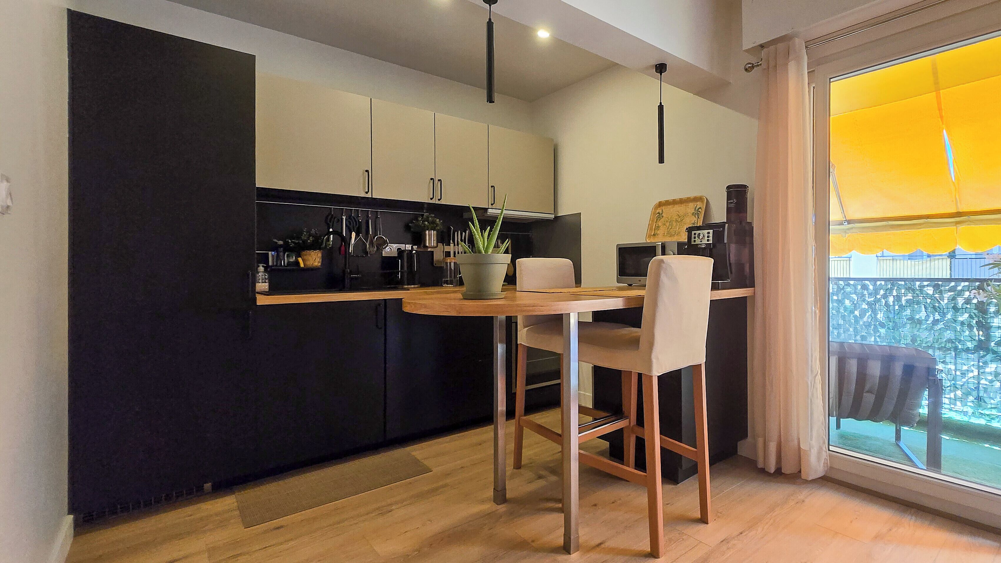 Eine moderne Küche mit dunklen Schränken, einem Holztisch und Blick auf ein gelbes Gebäude durch ein Fenster.