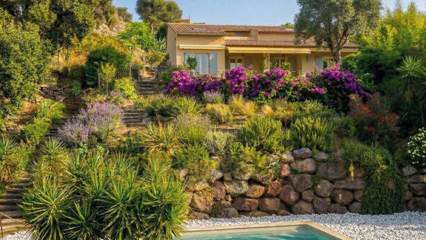 Ein Haus im mediterranen Stil mit Pool, umgeben von üppiger Vegetation und einer Steinmauer.
