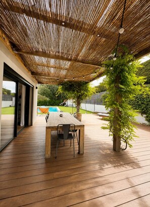 Eine Terrasse im Freien mit einem Holztisch und -stühlen, einem Reetdach und Blick auf einen Swimmingpool.