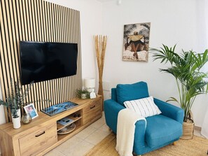 Ein Wohnzimmer mit Fernseher, einem blauen Sofa, einem Holz-TV-Schrank, einer Pflanze und einem Wandbild.