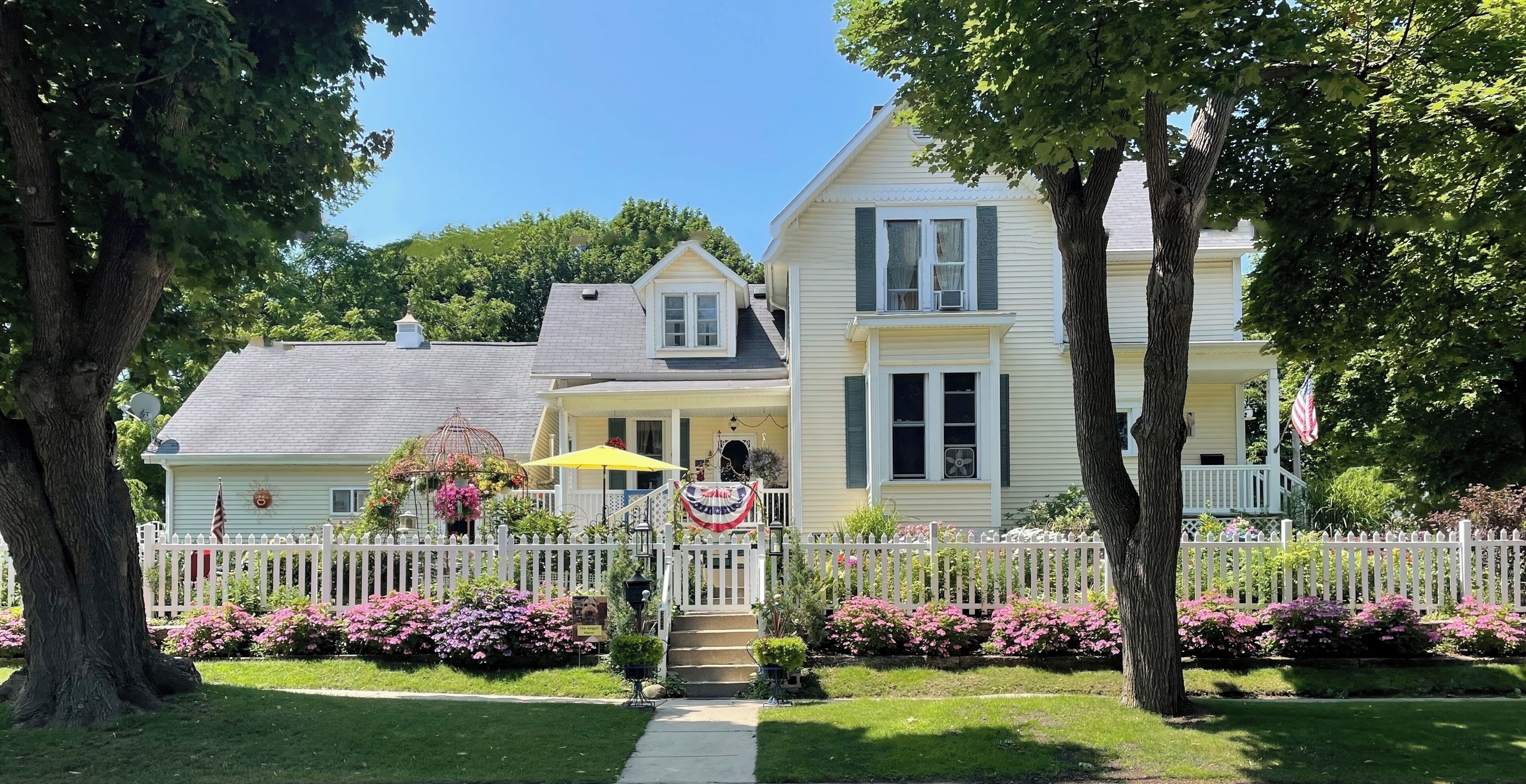 A two-story house with a white picket fence, a front porch, and a flag.