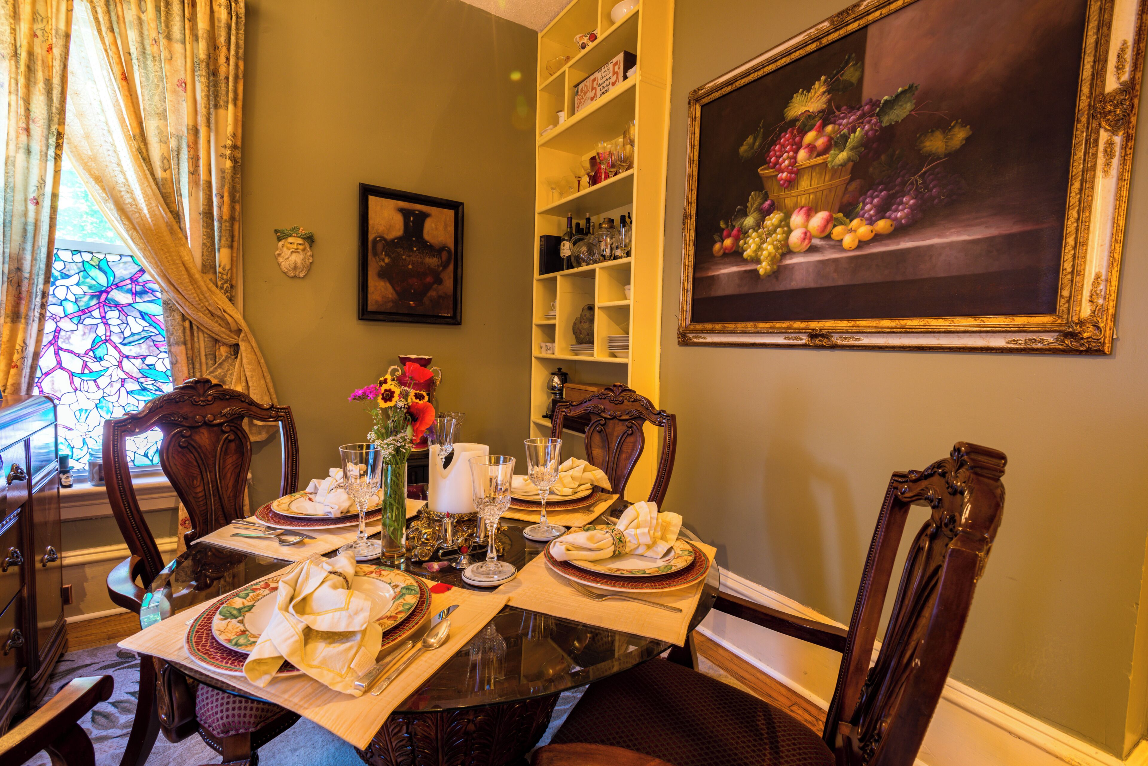 A dining room with a table set for four, surrounded by chairs, a stained glass window, and a painting of fruit.