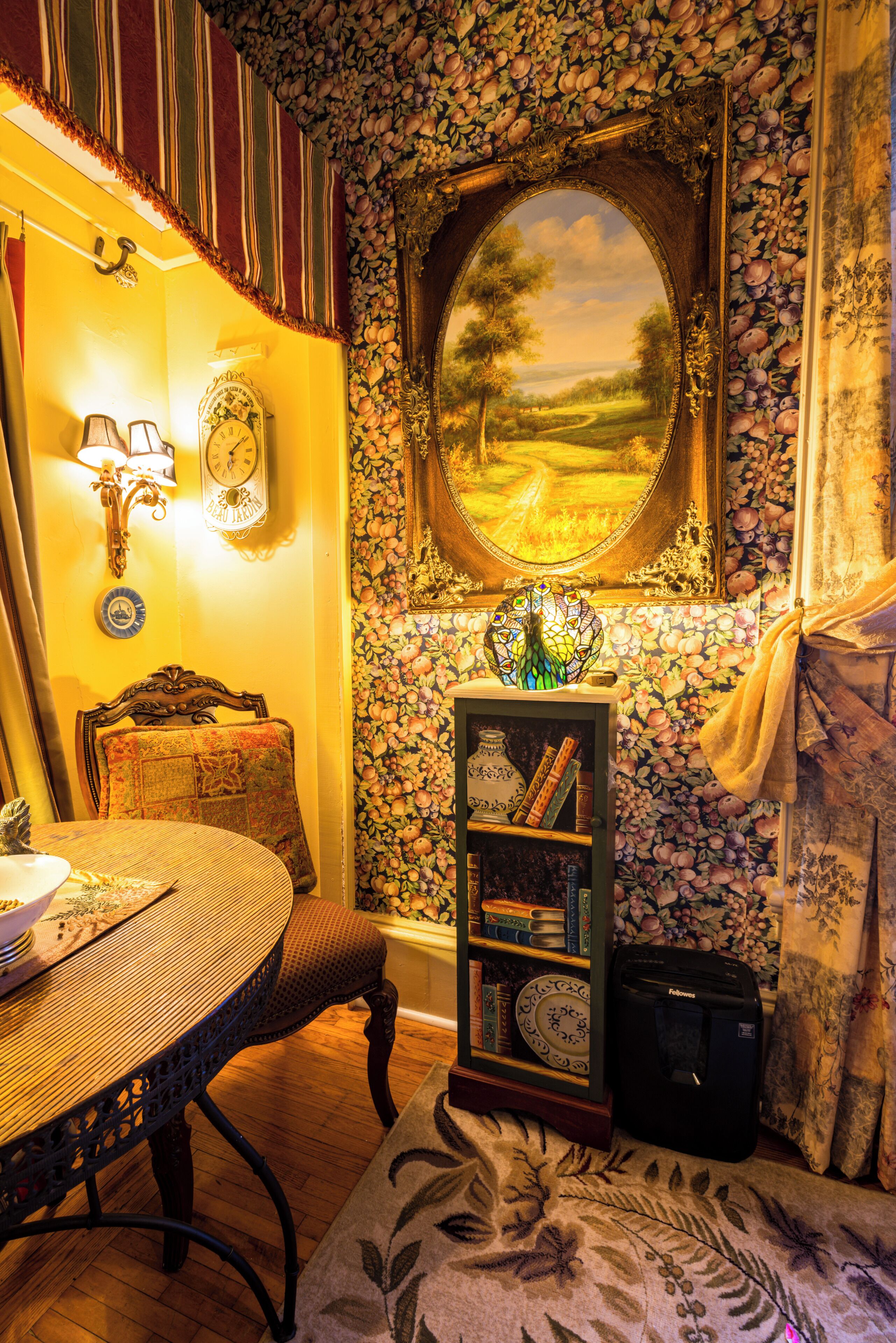 A room with a round table, a bookshelf, a clock, and a floral wallpaper.