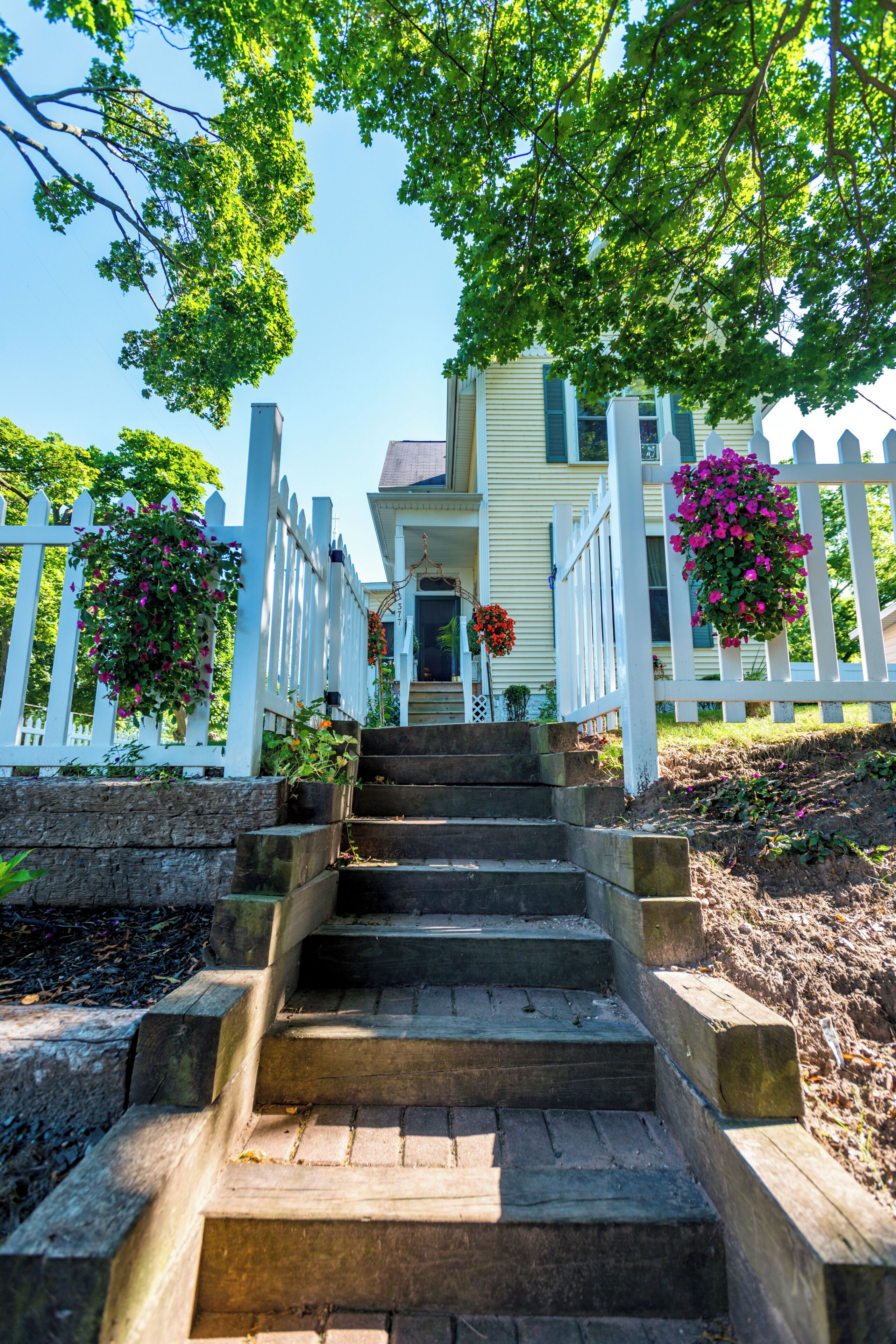 A set of stone steps leading to a house with a white picket fence, surrounded by greenery and hanging flower baskets.
