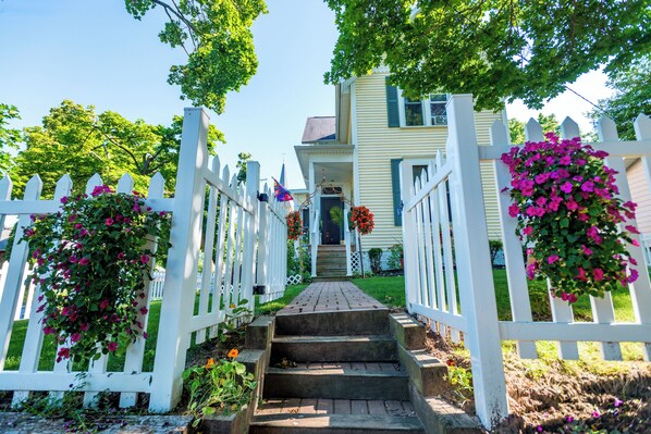 A house with a white picket fence, a brick pathway leading to the front door, and hanging flower baskets.