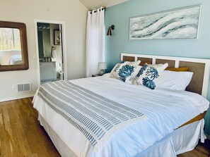 Ein Schlafzimmer mit Bett, Spiegel, Tür, Wandbild und einem Fenster mit Vorhängen.