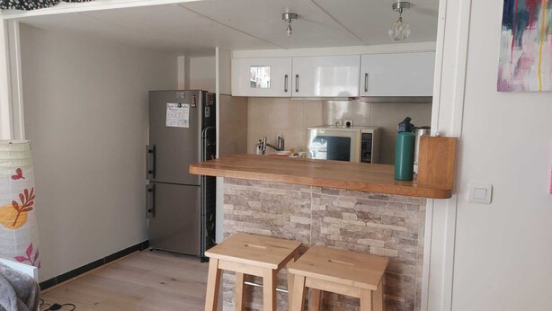 Cocina compacta con refrigerador, microondas y una pequeña encimera. Hay dos taburetes de madera y una botella de agua sobre la encimera.