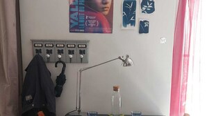 Una habitación con una mesa, dos sillas de madera, una lámpara, una botella y diversas obras de arte en la pared.