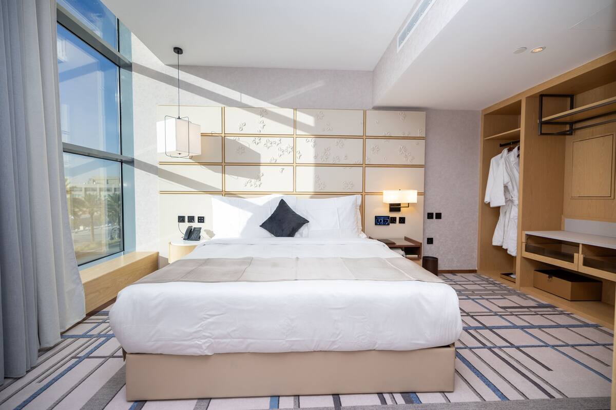 A modern hotel room with a large bed, a bedside table, a wardrobe, and a view through a large window.