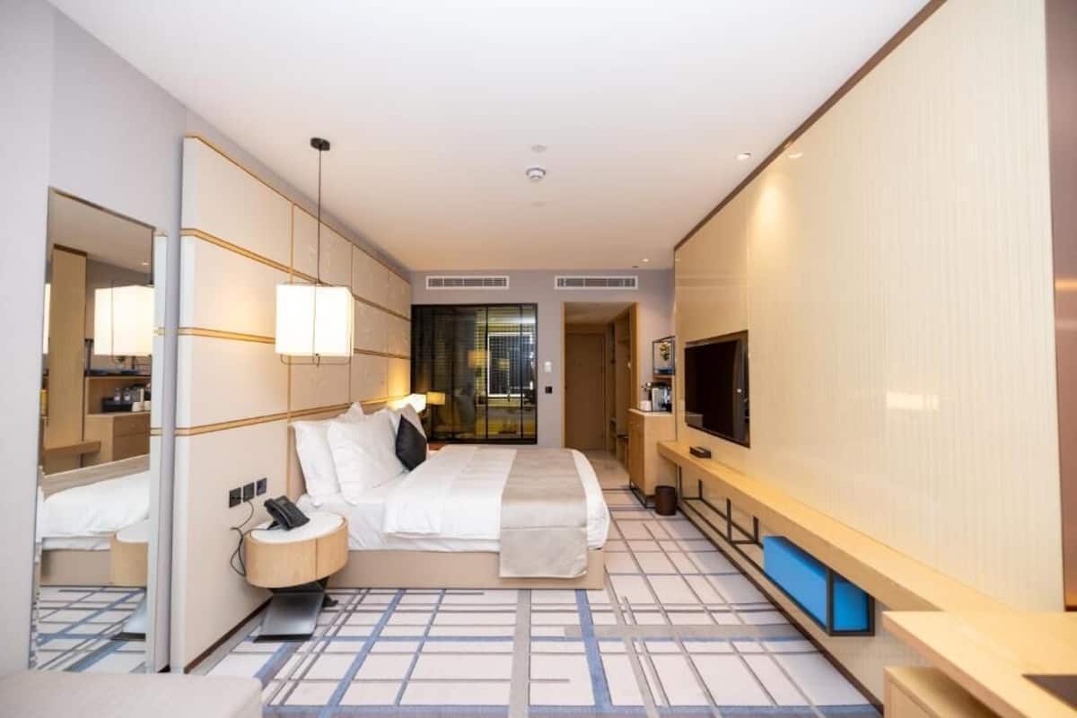 A hotel room with a bed, bedside table, lamp, TV, and a large mirror.