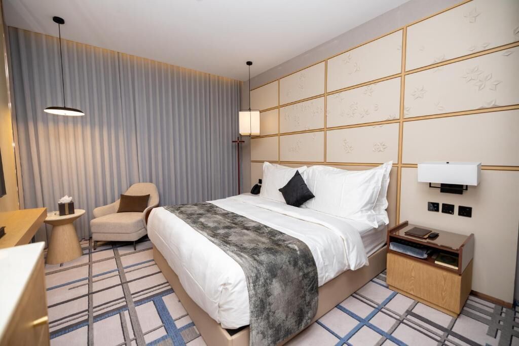 Een hotelkamer met een bed, nachtkastje, stoel en een groot raam met gordijnen.