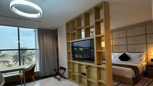 A modern hotel room with a large TV, a bed, a desk, and a view of palm trees. - 7Crown Tahliah Hotel (Riyadh)