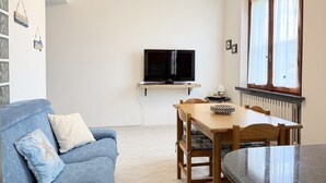 Een woonkamer met een bank, een eettafel, stoelen, een televisie op een dressoir, en een raam met gordijnen.