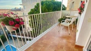 A balcony with a tiled floor, white railing, potted plants, and a view of buildings and trees. - Cosy aparment 200 meters from the beach (Vilanova i la Geltrú)