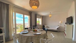 A dining area with a table set for two, a sofa, a TV, and a balcony with a view. - Cosy aparment 200 meters from the beach (Vilanova i la Geltrú)
