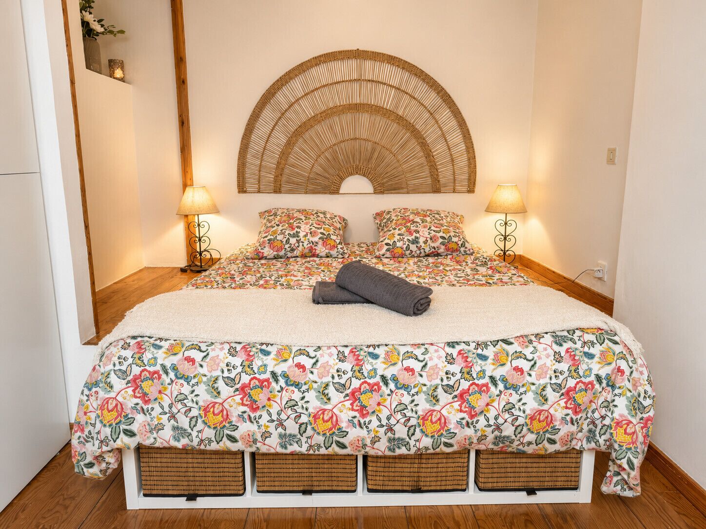A bed with a floral bedspread, two pillows, a rolled towel, a woven headboard, and two bedside lamps.