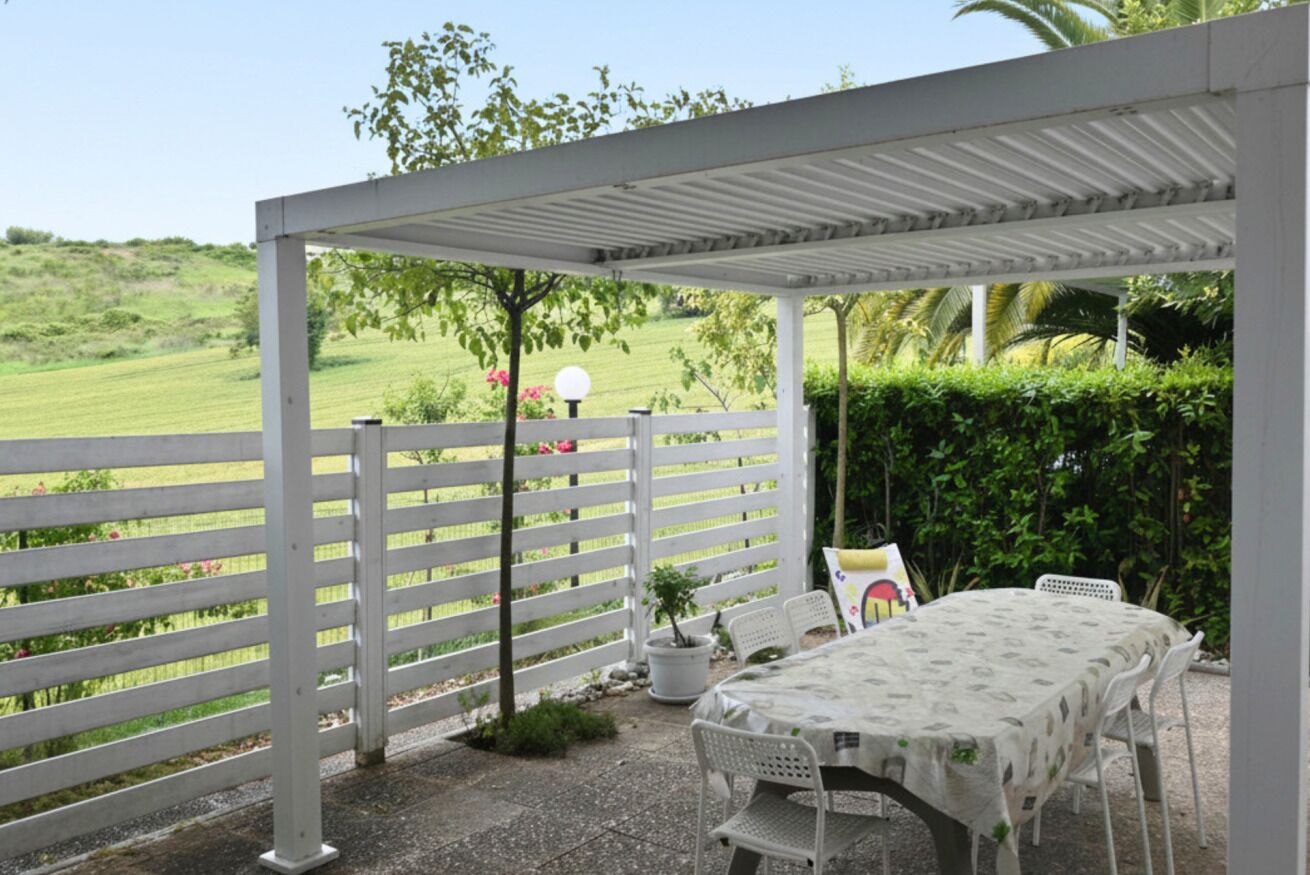 Ein überdachter Patio-Bereich mit Tisch und Stühlen, ein weißer Zaun und Blick auf eine grüne Landschaft.