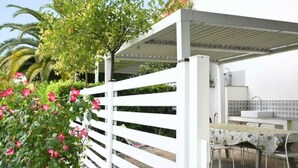 Une terrasse extérieure blanche, avec une table et des chaises, entourée de verdure et d’une clôture blanche.