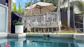 Et område ved poolen med en makrame-stol, en potteplante og en stribet parasol.