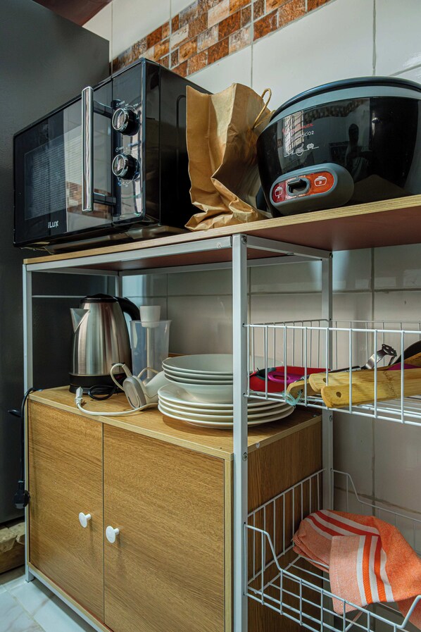 A kitchen with a microwave, kettle, and a shelf with plates and a cutting board.