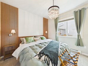 Ein Schlafzimmer mit einem Bett, Nachttischlampen, einer Wand aus Holzpaneelen, einem Fenster mit Vorhängen und einer hängenden Chandelier.