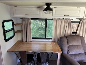 Un intĂ©rieur de camping-car compact comprenant une table Ă manger, deux chaises, une fenĂȘtre avec des rideaux et un luminaire au plafond.