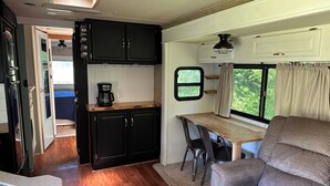 Cocina compacta para autocaravana con cafetera, zona de comedor con mesa y sillas, y vista por una ventana.