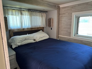 Un lit simple avec une tĂȘte de lit en bois, une table de chevet dotĂ©e dâune prise Ă©lectrique, une fenĂȘtre avec des stores et une Ă©tagĂšre fixĂ©e au mur.