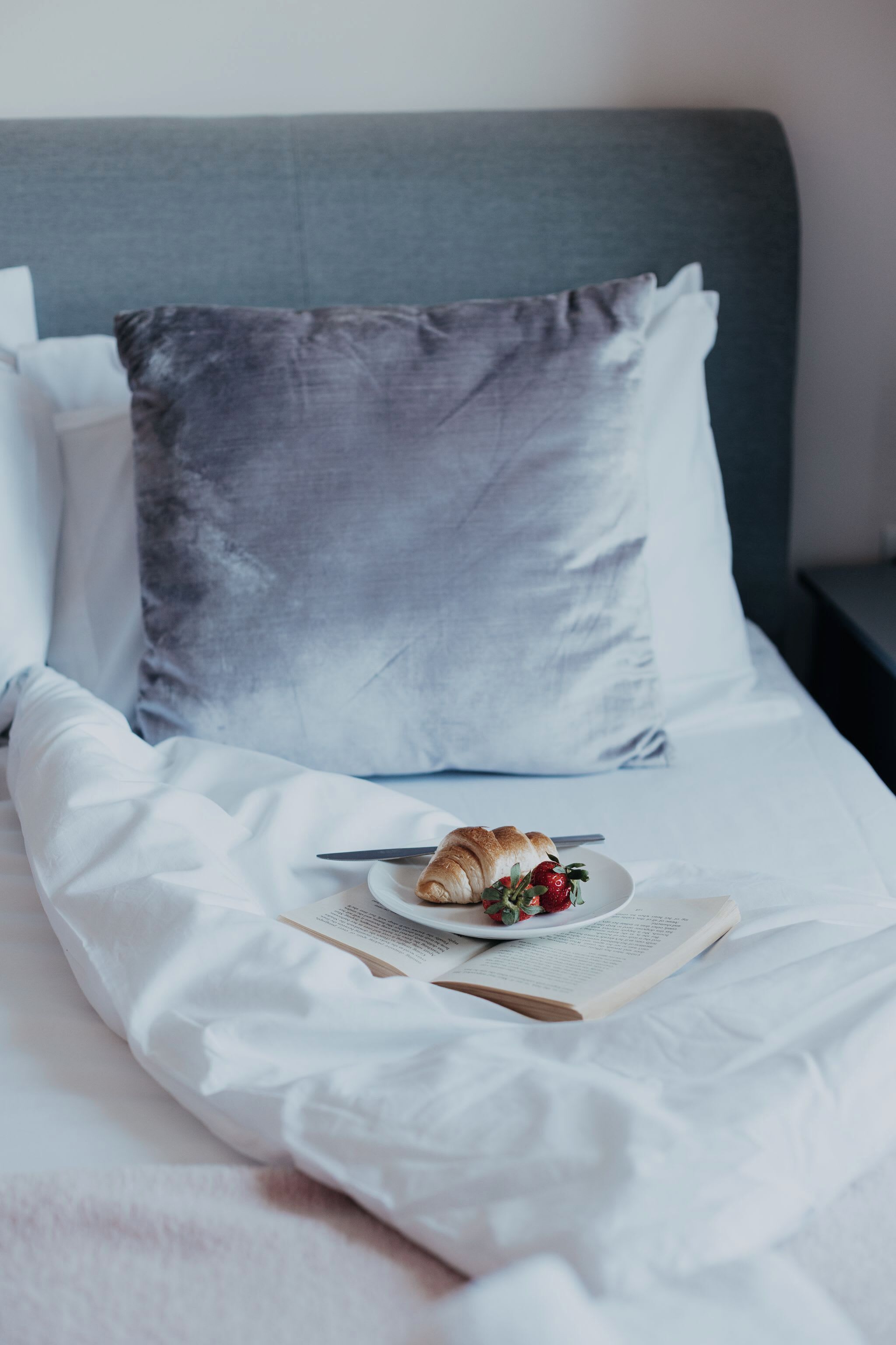 Cama bem arrumada com lençóis brancos, um travesseiro cinza, um prato com um croissant e morangos, e um livro aberto.