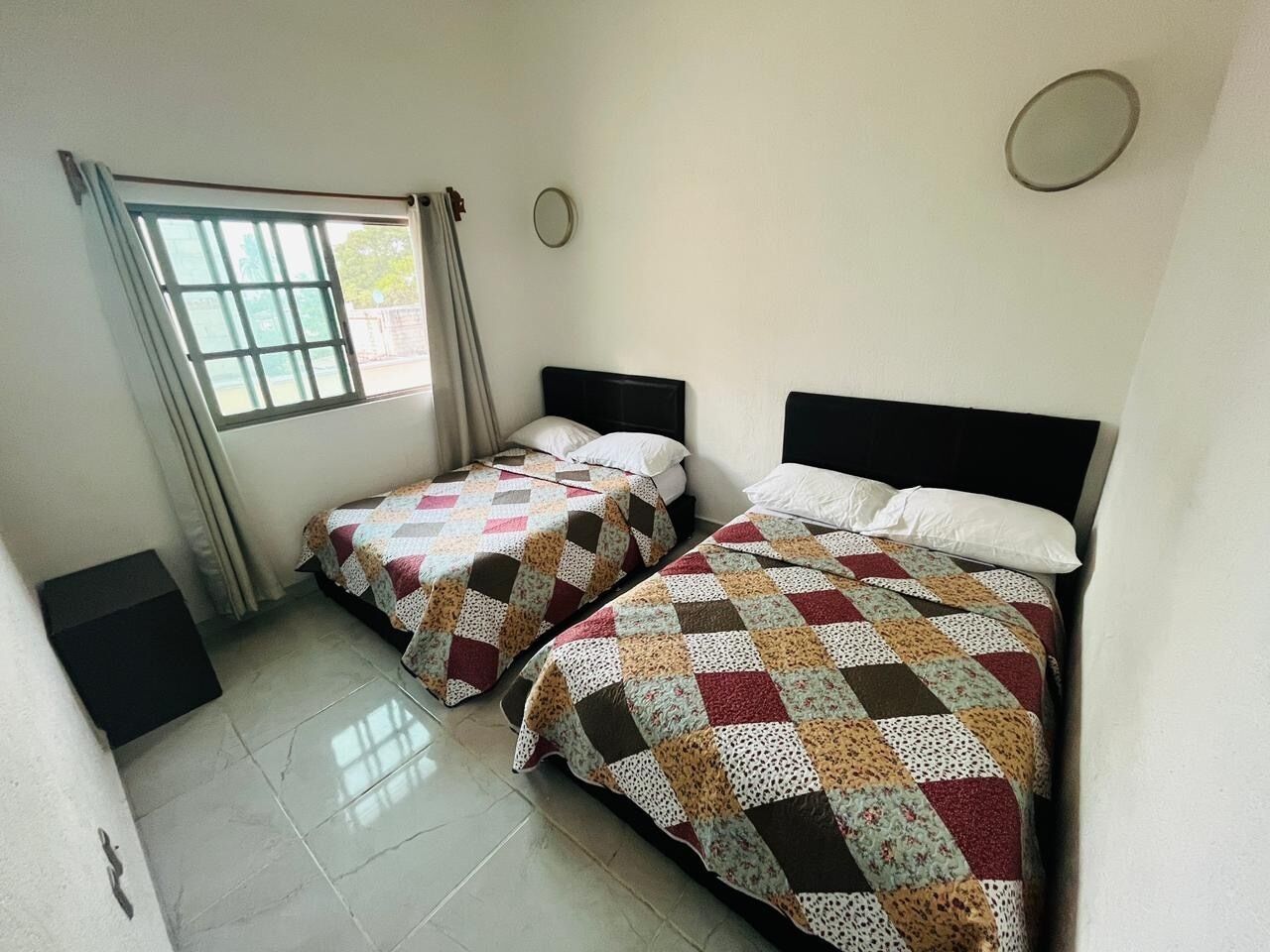 A room with two beds, a window with curtains, a round mirror, and a tiled floor.