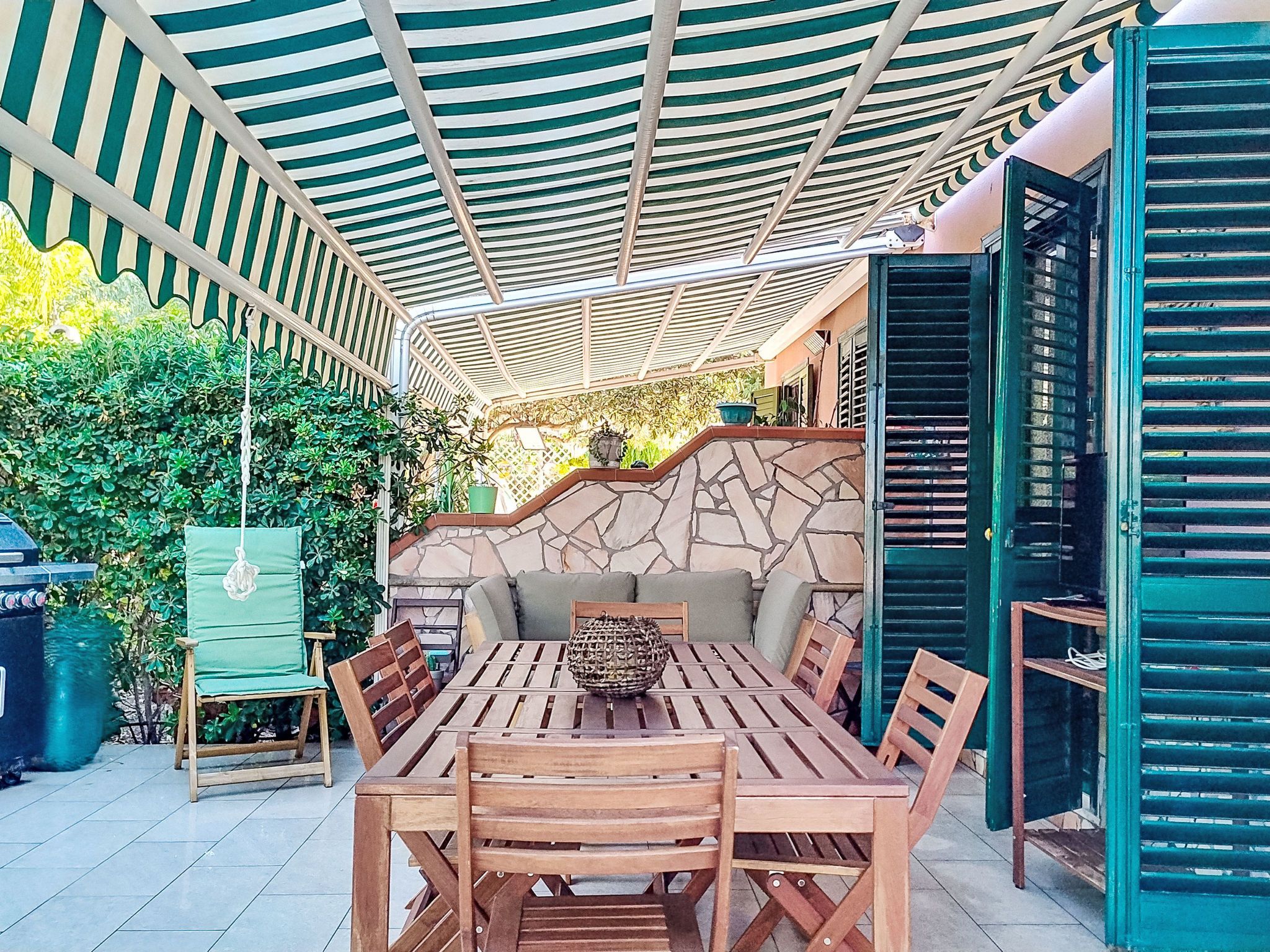 An outdoor patio with a stone wall, wooden furniture, and a covered area with a striped ceiling.