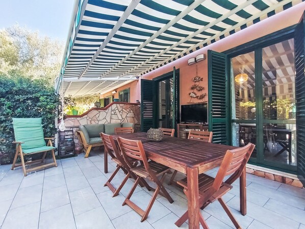 An outdoor patio with a wooden table and chairs, a covered seating area, and a stone wall.
