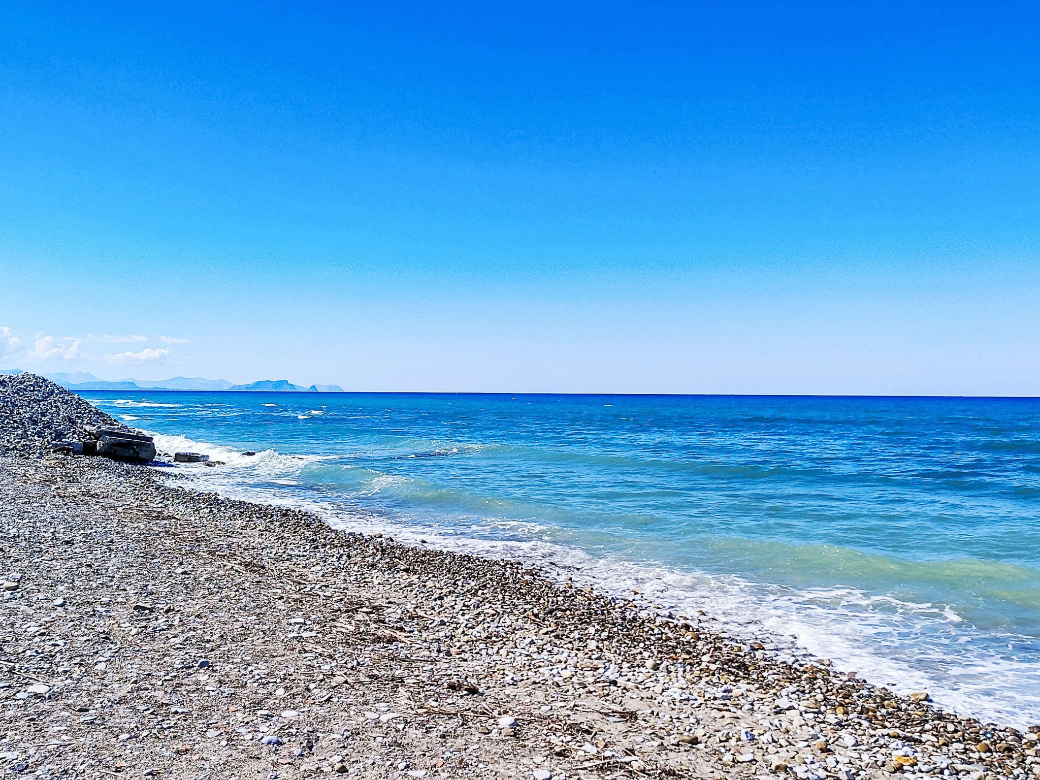 Une plage de galets bordée d’une eau d’un bleu limpide, avec des montagnes qui se dessinent à l’horizon.