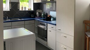 A compact kitchen with a built-in oven, a stove, and a refrigerator. There is a window providing natural light, and a small dining area with a chair. - Top location! Apartment at the foot of the Rhön, forest on the doorstep. Pet friendly! (Dipperz)