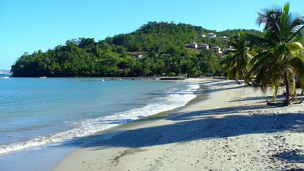 Una playa con vista despejada al mar, una orilla arenosa y una colina con edificios al fondo.